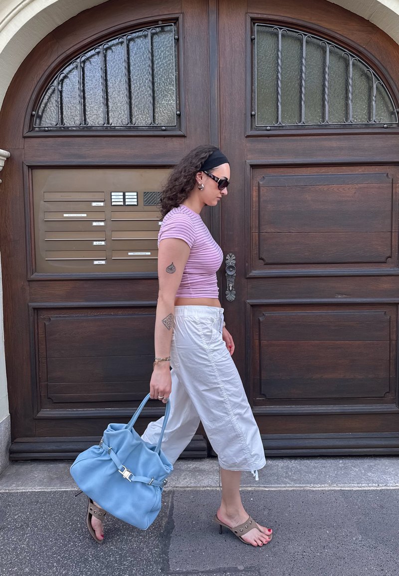 Femme portant des lunettes de soleil, top violet rayé, pantalon blanc, portant un grand sac bleu, marchant devant une porte en bois avec des boîtes aux lettres.