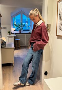 Bordowa, oversizeowa koszula z długim rękawem i białym kołnierzykiem, niebieskie jeansy o prostych nogawkach oraz beżowe sneakersy z bordowymi akcentami. Wnętrze.