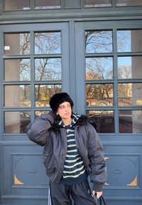 Sort bomberjakke med kunstpelskant, lagd over en stripete hettegenser. Mørke bukser og svart pelslue, holder en håndveske, foran vinduer.