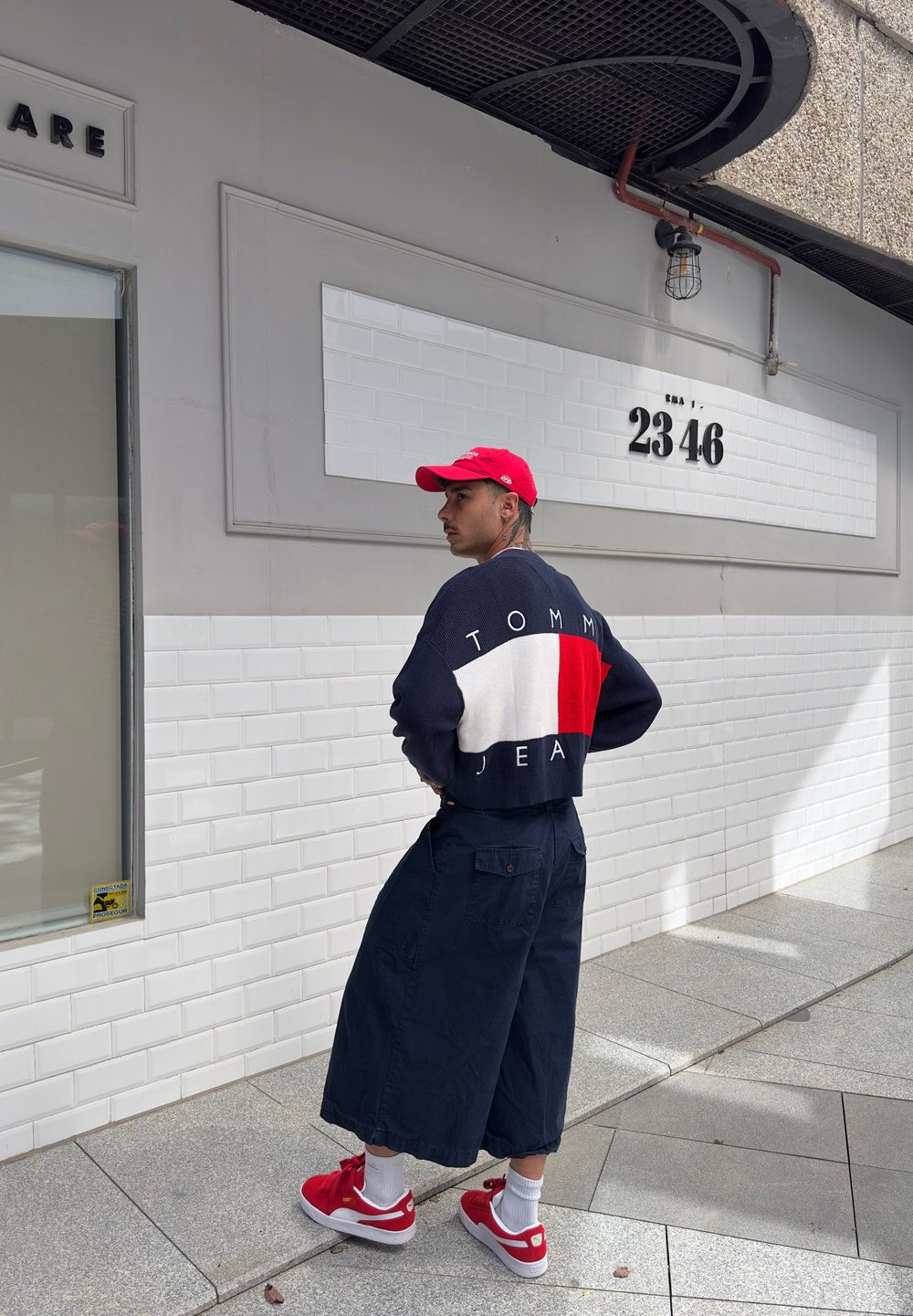 Ναυτικό και κόκκινο πλεκτό πουλόβερ με άσπρη λωρίδα, με την ετικέτα "TOMMY JEAN", συνδυασμένο με φαρδιές ναυτικές βερμούδες και κόκκινες αθλητικές παπούτσια με λευκές κάλτσες.