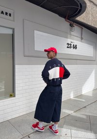 Ναυτικό και κόκκινο πλεκτό πουλόβερ με άσπρη λωρίδα, με την ετικέτα "TOMMY JEAN", συνδυασμένο με φαρδιές ναυτικές βερμούδες και κόκκινες αθλητικές παπούτσια με λευκές κάλτσες.