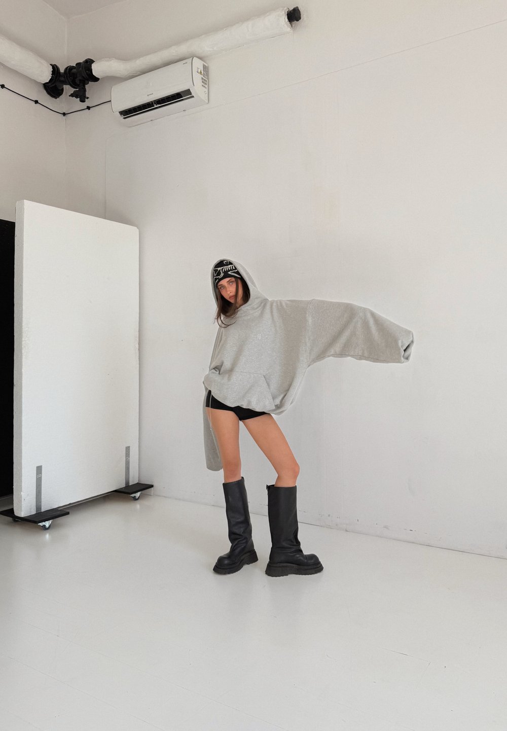 Přehnaně velká šedá mikina s kapucí a předním kapsou. Kombinována s černými vysokými kozačkami nad kolena. Jednoduché pozadí se studiovým osvětlením.