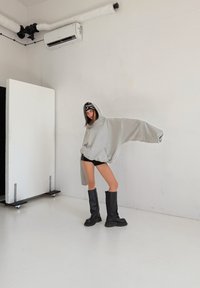 Přehnaně velká šedá mikina s kapucí a předním kapsou. Kombinována s černými vysokými kozačkami nad kolena. Jednoduché pozadí se studiovým osvětlením.