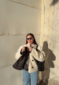 Femme portant des lunettes de soleil, une veste en peau lainée couleur crème, un pull à rayures et un jean bleu, debout contre un mur beige texturé au soleil.