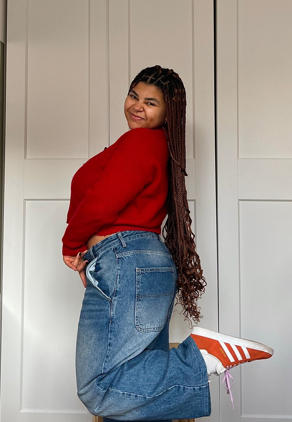 Κόκκινο ριγέ πουλόβερ, ψηλόμεσα μπλε τζιν με φθαρμένες λεπτομέρειες και πορτοκαλί και λευκά αθλητικά παπούτσια, ποζάροντας μπροστά σε λευκό φόντο.