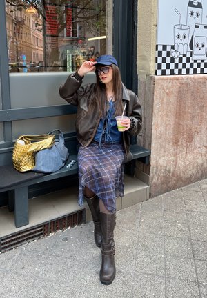 Mujer con chaqueta de cuero marrón y falda de cuadros sentado en un banco fuera de una cafetería, sosteniendo una bebida verde con pajita, ajustándose una gorra azul marino.