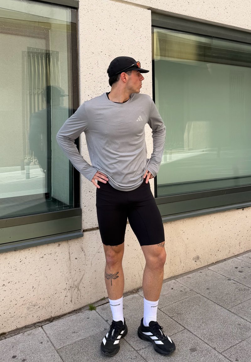 Férfi fekete sapkában, szürke hosszú ujjú ingben, fekete rövidnadrágban és sportcipőben áll, kezeit a csípőjén tartva, oldalra nézve az épület ablakai közelében.