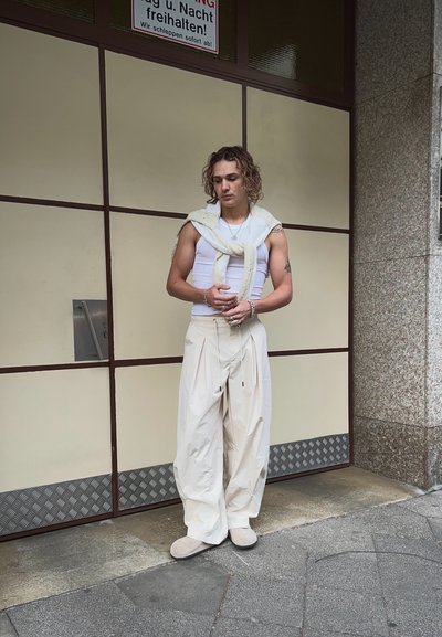 Modelo vistiendo una camiseta sin mangas blanca, pantalones anchos color crema y zapatillas beige, con un suéter texturizado beige caído sobre los hombros.