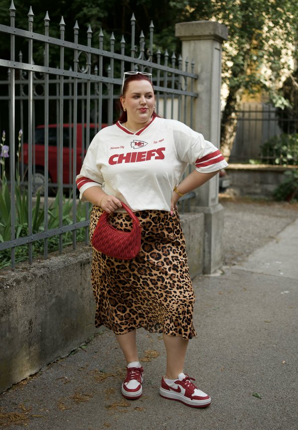 Balta marškinėliai su raudonu "CHIEFS" užrašu, raudonai juostuoti rankovės, leopardo rašto midi sijonas, raudonas pintas rankinukas ir raudoni/balti sportbačiai.