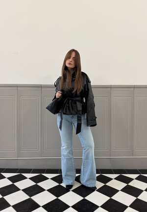 Mujer con cabello largo, vestida con una chaqueta negra de cuero con cinturón, vaqueros acampanados azul claro y de pie sobre un suelo a cuadros en blanco y negro.