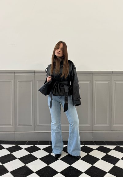 Mujer con cabello largo, vestida con una chaqueta negra de cuero con cinturón, vaqueros acampanados azul claro y de pie sobre un suelo a cuadros en blanco y negro.