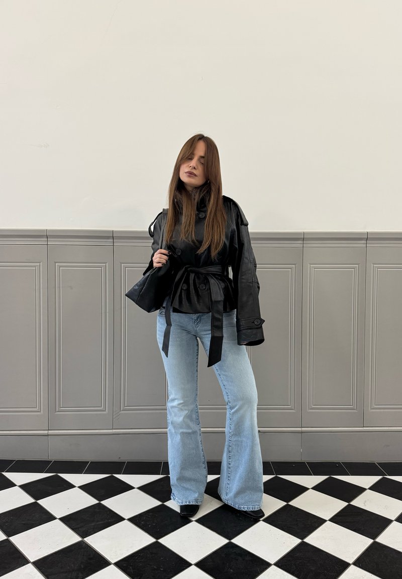 Femme aux longs cheveux portant une veste en cuir noire ceinturée, un jean évasé bleu clair, debout sur un sol en damier noir et blanc.