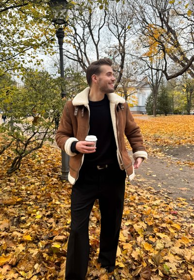 Chaqueta de piel de oveja marrón con forro crema, suéter negro y pantalones negros, sosteniendo una taza de café, rodeado de hojas de otoño en un parque.