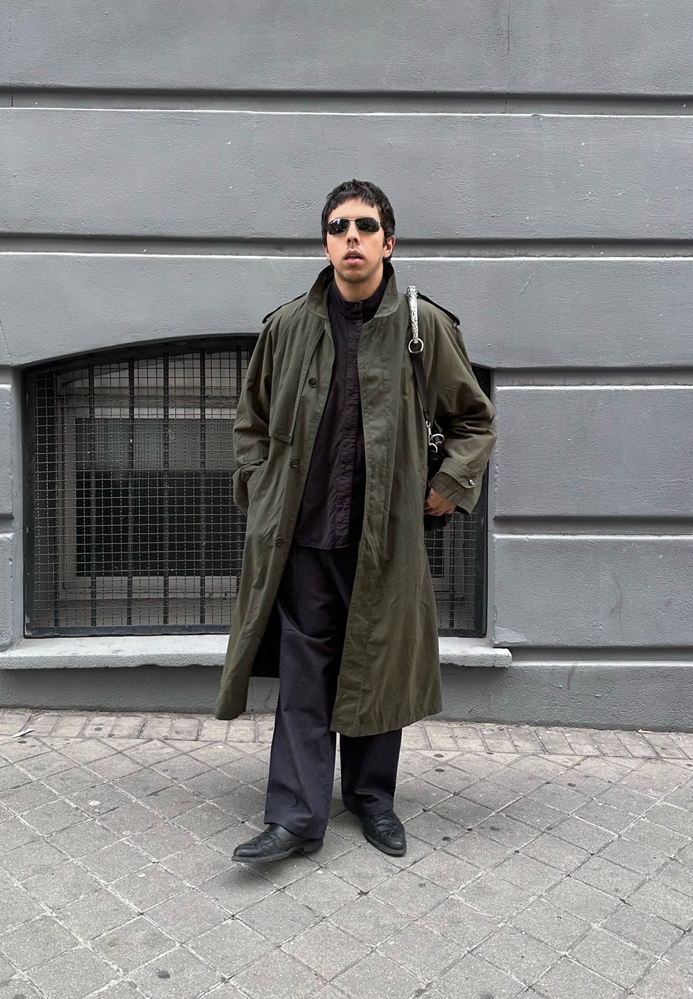 Homme portant des lunettes de soleil noires, un long manteau vert, un pantalon noir et des chaussures, debout les mains dans les poches sur un trottoir urbain devant un mur gris avec une fenêtre à barreaux.