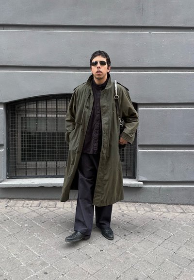 Hombre con gafas de sol oscuras, abrigo largo verde, pantalones y zapatos negros, de pie con las manos en los bolsillos en una acera urbana frente a una pared gris con ventana enrejada.