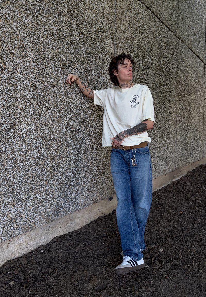Personne avec des tatouages portant un t-shirt blanc Adidas et un jean bleu, appuyée contre un mur de pierre texturé sur un sol en terre, regardant sur le côté.