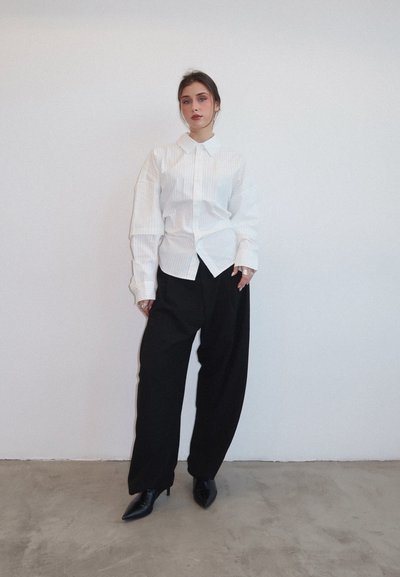 Camisa a rayas blancas con cuello puntiagudo, de corte oversize, combinada con pantalones negros holgados y tacones negros puntiagudos, sobre un fondo liso.
