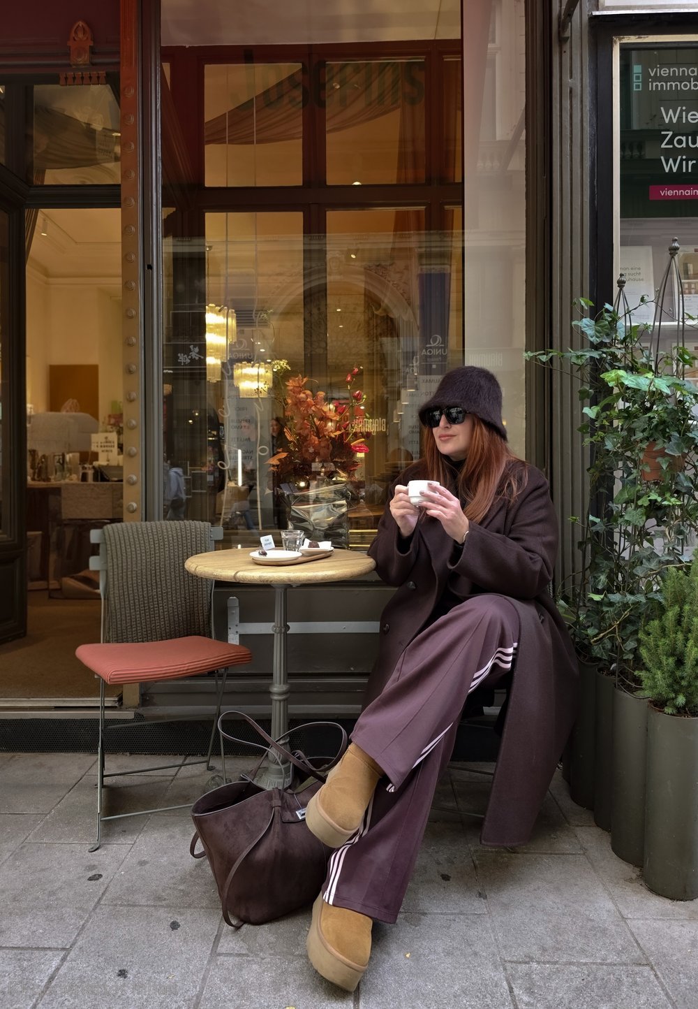 Manteau et pantalon marron avec des accents rayés, bottes en suède marron, assis à une table circulaire avec un plateau en bois clair, chaises grises et rouges, cadre urbain.