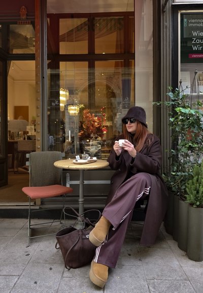 Abrigo y pantalones marrón con acentos a rayas, botas de ante marrón, sentado en una mesa circular con tapa de madera clara, sillas grises y rojas, entorno urbano.