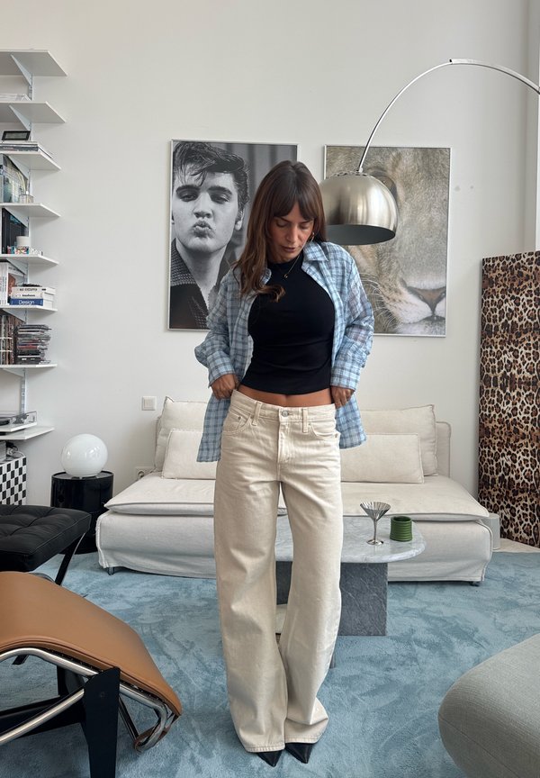 Una mujer está de pie en un salón y se ajusta unos pantalones color crema de pernera ancha sobre un top negro y una camisa oversize de cuadros azules.