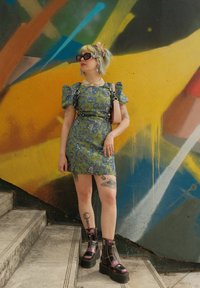 Πράσινο φλοράλ φόρεμα με φουσκωτά μανίκια, σε συνδυασμό με μαύρες πλατφόρμες. Αξεσουάρ περιλαμβάνουν μια κοσμητική χάντρα και γυαλιά ηλίου. Ένα πολύχρωμο γκράφιτι στο φόντο.