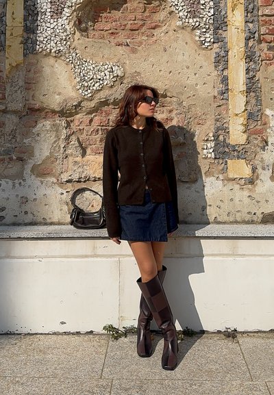 Cardigan de lana marrón con botones, combinado con una minifalda de mezclilla y botas marrones de tacón cuadrado hasta la rodilla; bolso de hombro negro. Fondo con textura en la pared.