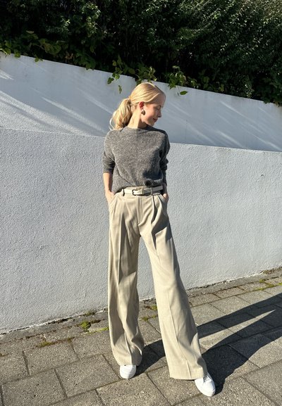 Mujer con coleta rubia, vistiendo un suéter gris, pantalones de pierna ancha beige y zapatillas blancas, se encuentra contra una pared blanca con vegetación arriba.