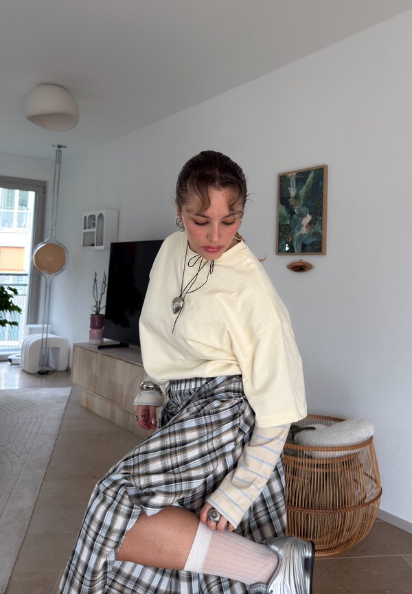 Une femme portant un sweat-shirt crème et une jupe à carreaux est assise dans un salon et ajuste sa chaussette rayée au-dessus d’une basket argentée.