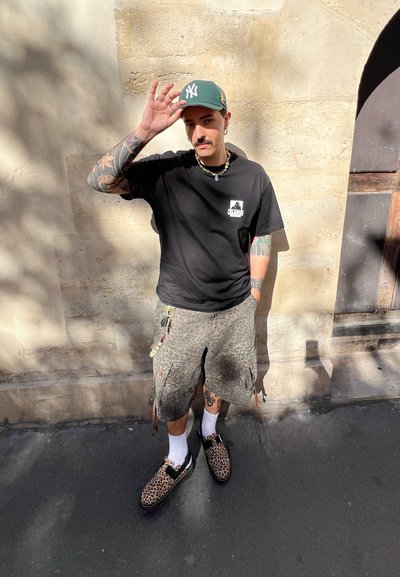 Hombre con gorra verde, camiseta negra, pantalones cortos de estampado de leopardo y zapatos se apoya contra una pared beige, levantando una mano hacia la gorra.