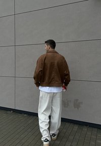 Veste en daim marron avec un col, t-shirt blanc en dessous, et pantalon en velours côtelé de couleur claire. Baskets noires avec des accents blancs.