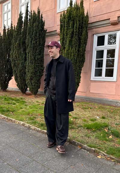 Abrigo negro sobre una camiseta negra, pantalones negros holgados y zapatos marrones. Una gorra de béisbol color burdeos y arbustos verdes al fondo.