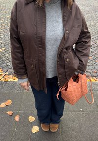 Giacca marrone con cerniera e due tasche frontali, indossata sopra un maglione grigio. Jeans blu scuro e una borsa intrecciata color pesca con trama.