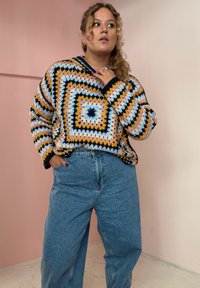 Suéter de crochê colorido com padrões geométricos em amarelo, azul e rosa, combinado com jeans de perna larga azul claro. Tecido texturizado, com capuz.