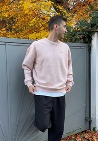 Lichtroze sweatshirt met geribbelde manchetten en zoom, gedragen over een wit shirt. Het model draagt een zwarte broek en staat bij een grijze poort.