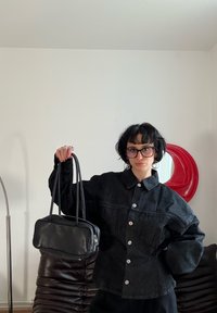 Personne aux cheveux courts et foncés portant des lunettes et une veste noire boutonnée, tenant un sac à main en cuir noir dans une pièce minimaliste.