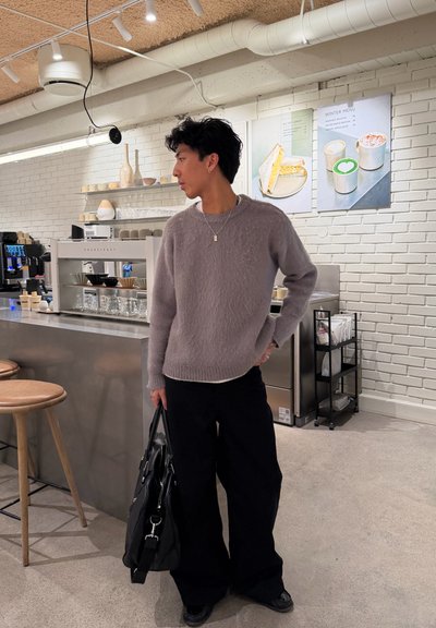 Joven con suéter gris y pantalones negros sosteniendo una bolsa, de pie dentro de una cafetería moderna cerca del mostrador y los letreros del menú.