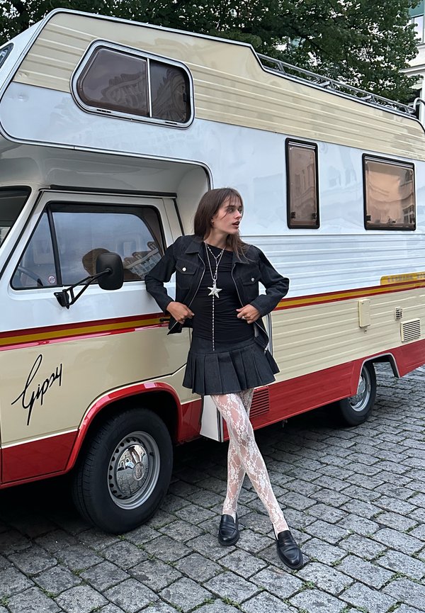 Czarna dżinsowa kurtka, czarna bluzka, plisowana czarna spódnica, białe koronkowe rajstopy i czarne buty. Vintage kamper w kolorze białym i beżowym w tle.