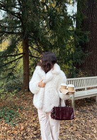 Bijeli kaput od umjetnog krzna s visokim ovratnikom, u kombinaciji sa svijetlo bež hlačama. Drži smeđu torbu s kavama i tamnocrvenu torbicu.