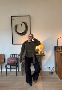 Femme portant des lunettes, une veste olive ceinturée et un pantalon noir, se tenant dans une pièce avec un sol en bois, une œuvre d'art murale abstraite et des meubles avec des lampes.