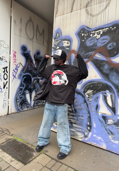 Camisa negra de gran tamaño con un diseño gráfico en rojo y blanco, combinada con jeans baggy de un azul claro y zapatos negros, frente a un fondo de graffiti.