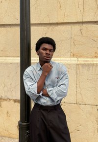 Dlhá modrá košeľa s jemnými vzormi, skombinovaná s čiernymi nohavicami s vysok�ým pásom. Osoba stojí opretá o textúrovanú stenu.