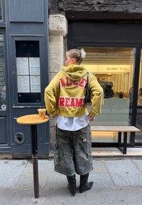 Sweat à capuche court jaune avec texte rouge "ALLIE DREAMS", assorti à un pantalon cargo camouflage et des bottines à talons noires, près d'une table en bois et d'un café.