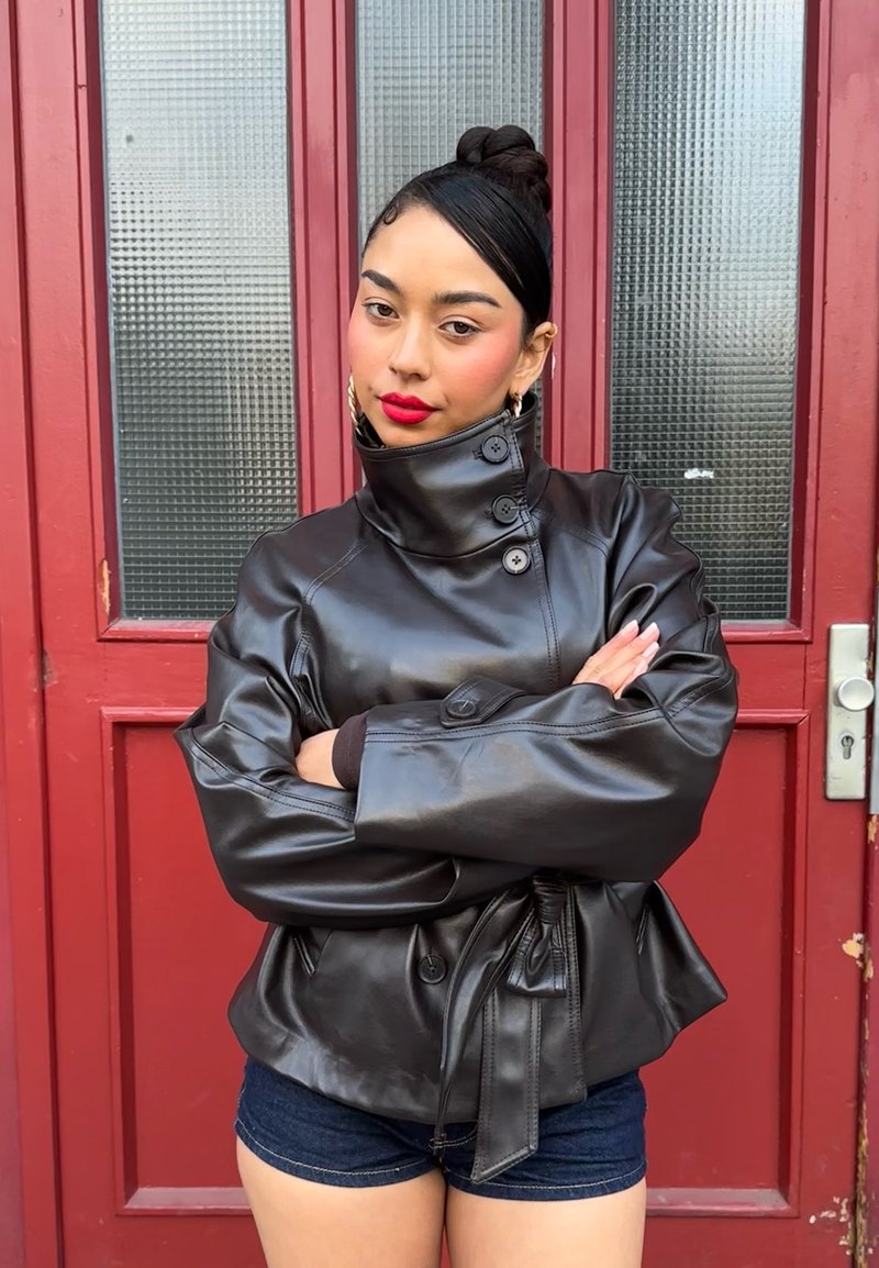 Femme aux cheveux foncés en chignon porte une veste en cuir noire boutonnée et un short en jean, se tenant avec confiance les bras croisés devant une porte rouge.