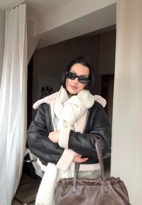 Geacă bomber neagră din piele cu guler de blană, eșarfă albă și geantă maro. Ochelari de soare întunecați și top deschis la culoare. Cadru interior.