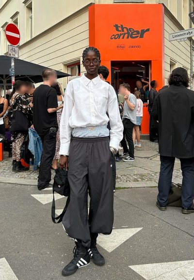 Pantalones deportivos negros con cintura ajustada, combinados con una camisa blanca de botones. El fondo presenta una colorida tienda de naranja brillante y una multitud.