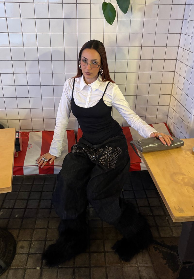Young woman with glasses sits between two tables on red and white cushion, wearing black wide pants with fluffy lower legs, layered top.