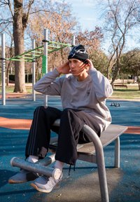 Sweat-shirt gris avec logo noir, pantalon en velours côtelé noir et bonnet noir avec un motif blanc. Assis sur un banc de musculation métallique dans un parc.