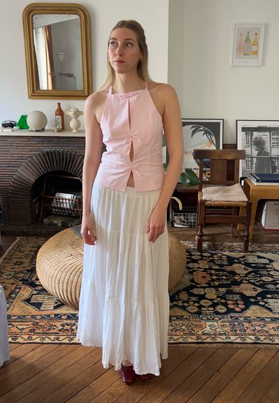Top sin mangas de color rosa claro con lazos, corte delantero y detalle de botones, combinado con una larga falda blanca de capas y zapatos de color rojo oscuro.