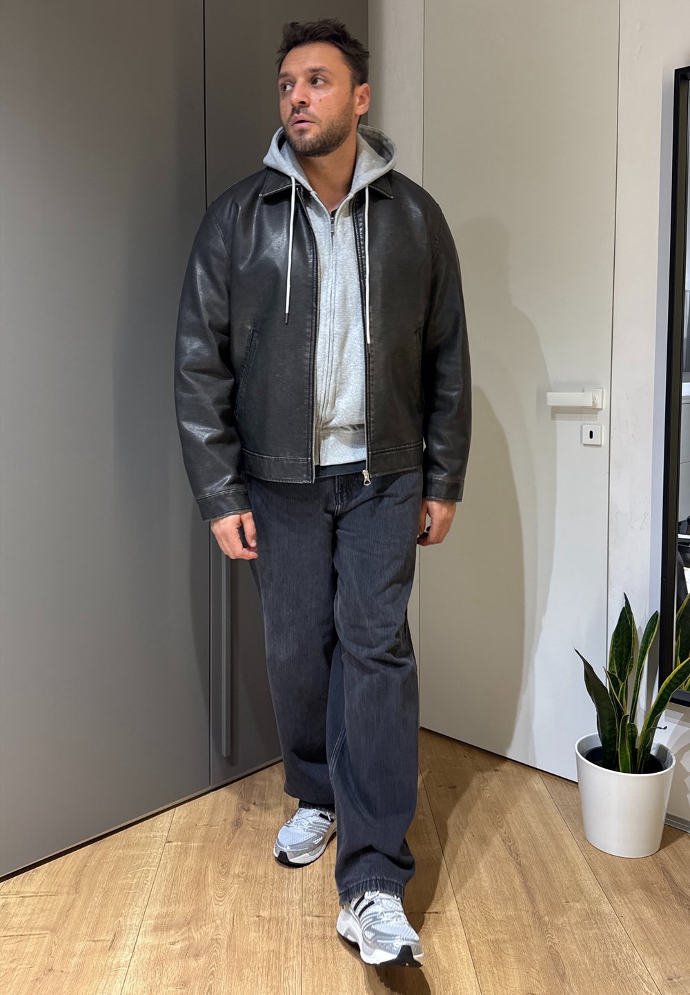 Veste en cuir noire sur un sweat à capuche gris, associée à un jean foncé et des baskets grises. Cadre intérieur avec une plante en pot dans un pot blanc.