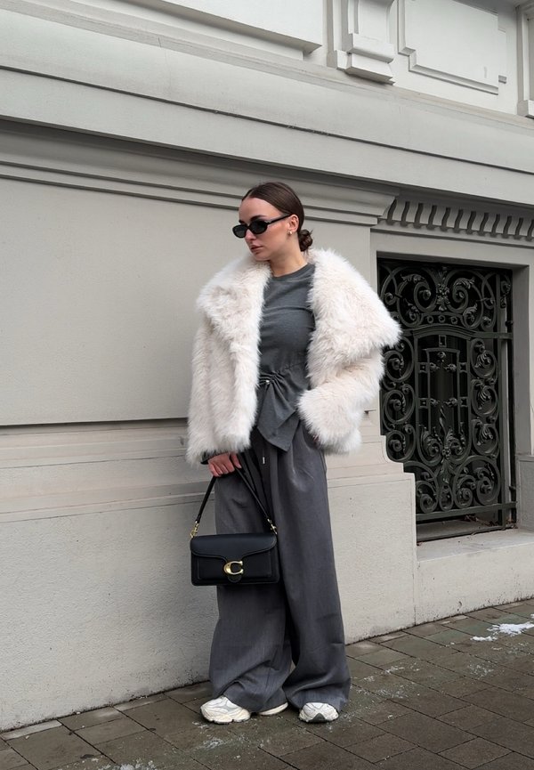 Donna che indossa un cappotto di pelliccia ecologica bianco, maglia grigia e pantaloni larghi, che tiene una borsa nera, in piedi vicino a un elaborato cancello di ferro nero su una strada cittadina.